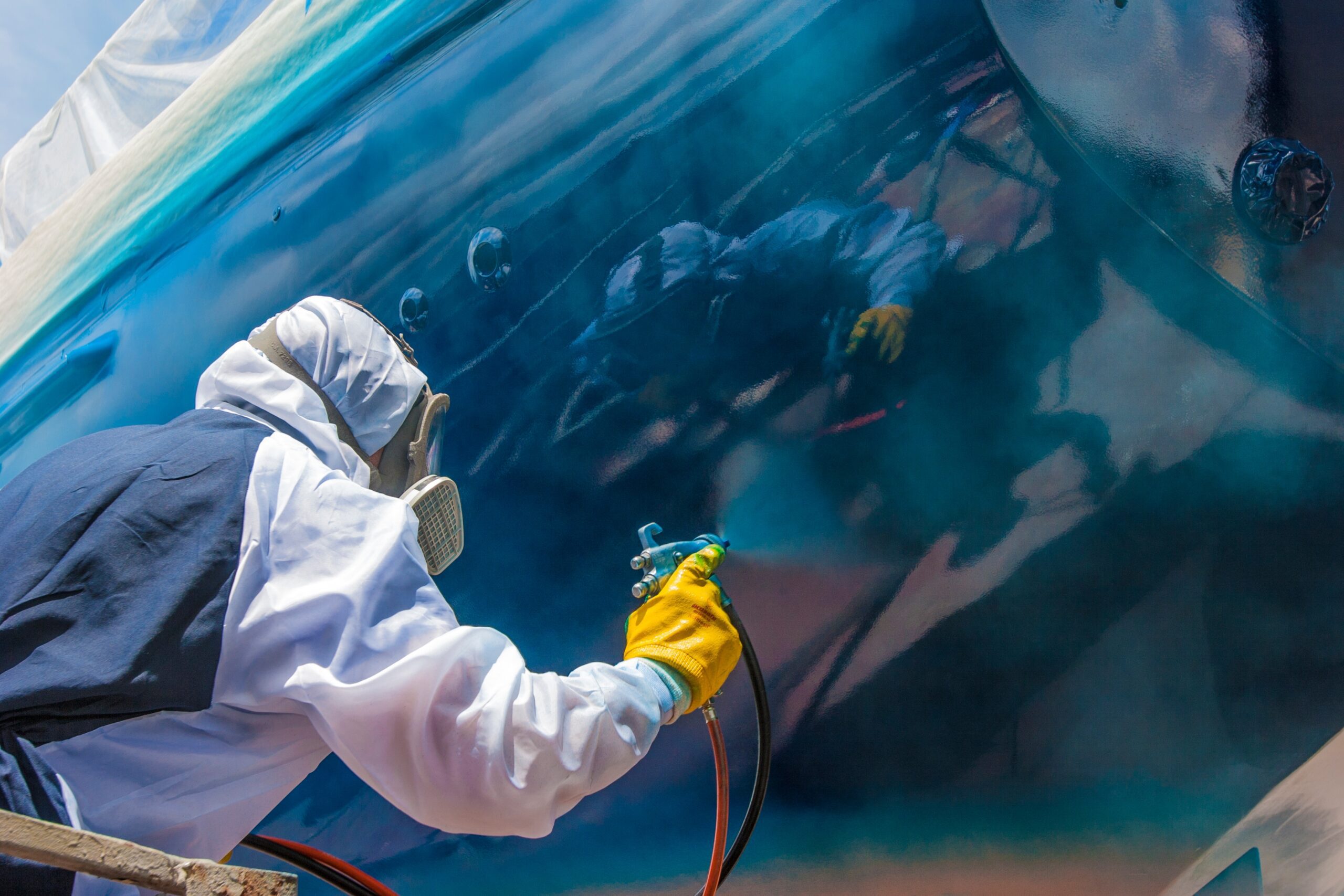 Spray painter with full body protection and mask painting the hull of sailboat with high gloss varnish